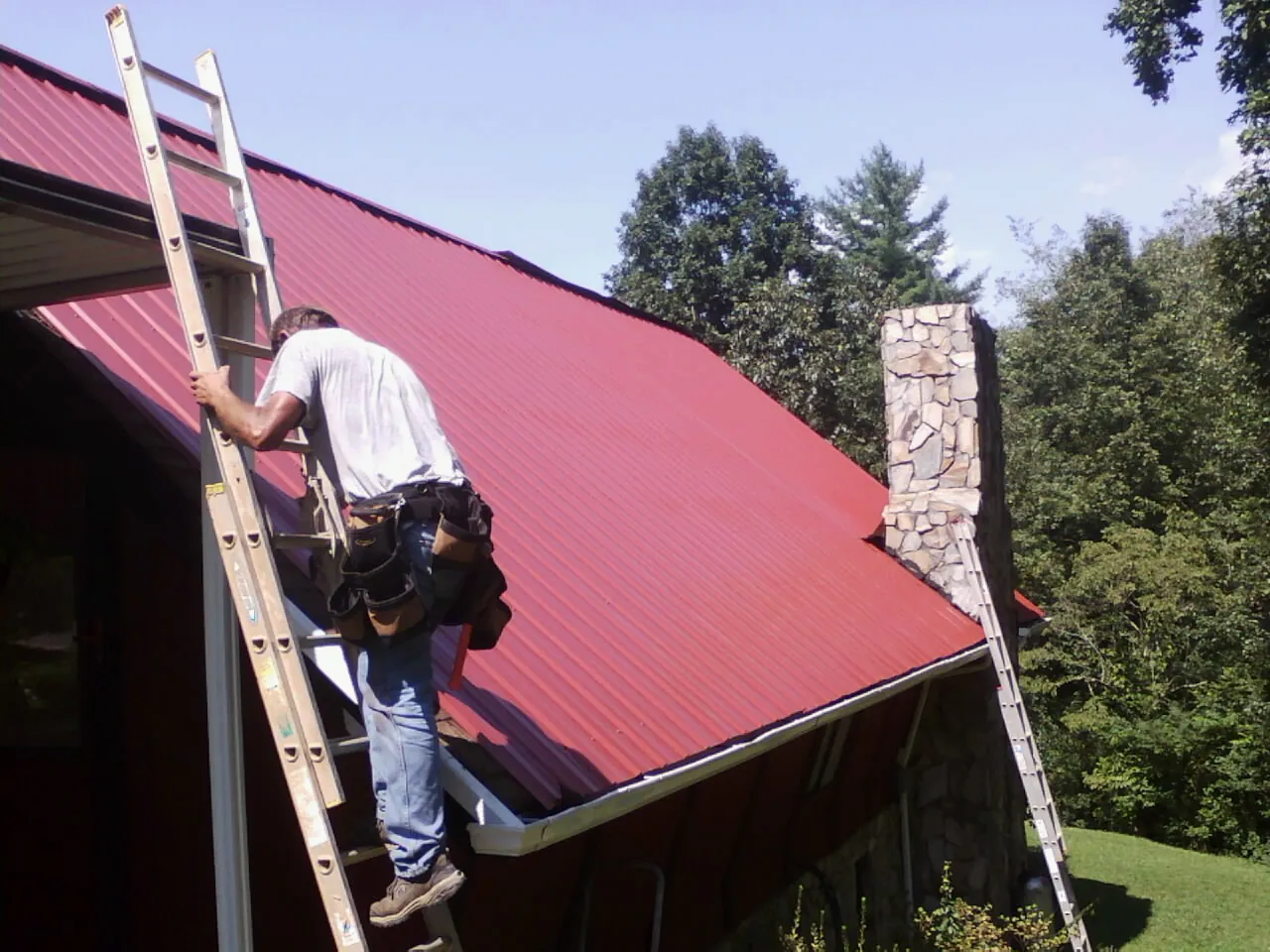 Roofer on a ladder beside a red metal roof and stone chimney — Soffit & Fascia Repair by Roofing Brothers in Osage Beach